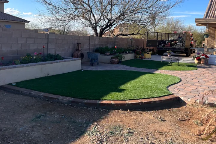 A small kidney-shaped patch of lush artificial grass framed by a red brick border and stone patio.